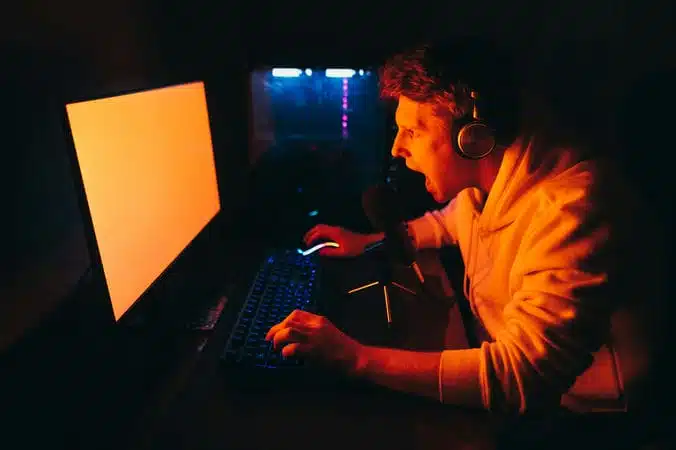 Portrait of emotional gamer in headphones playing computer games at home at table at night, yelling into microphone and looking intently at computer screen. Expressive gamer broadcasts a game stream
